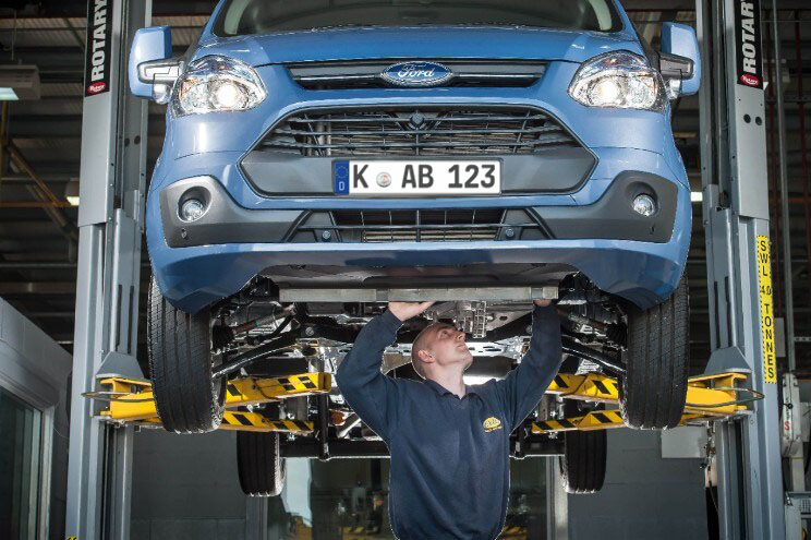 Ein Ford Transit steht in einer Werkstatt auf einem Wagenheber, während ein Mechaniker in blauer Arbeitskleidung darunter arbeitet. Das Bild zeigt eine typische Servicearbeit an dem Nutzfahrzeug.