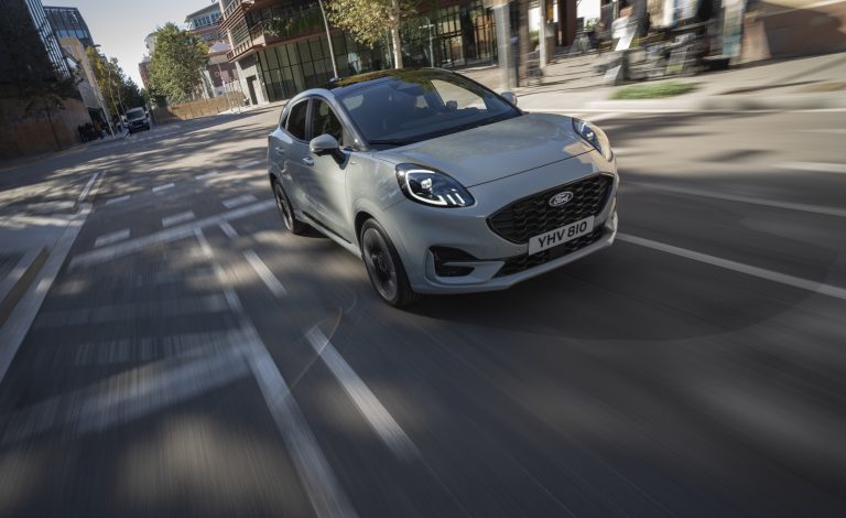 Ein Ford Puma fährt durch eine belebte Stadtstraße, vorbei an modernen Gebäuden. Das kompakte SUV zeigt sich agil im urbanen Verkehr, mit reflektierenden Scheiben und sportlicher Frontpartie.