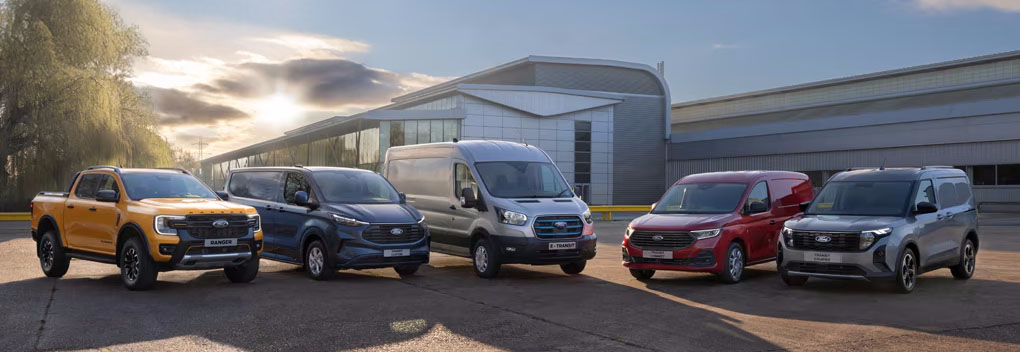Vier unterschiedliche Ford Transit-Modelle in verschiedenen Größen und Farben stehen gemeinsam mit einem robusten Ford Ranger vor eines modernen Gebäudes. Die Aufnahme präsentiert die ganze Bandbreite der Ford Nutzfahrzeug-Palette.