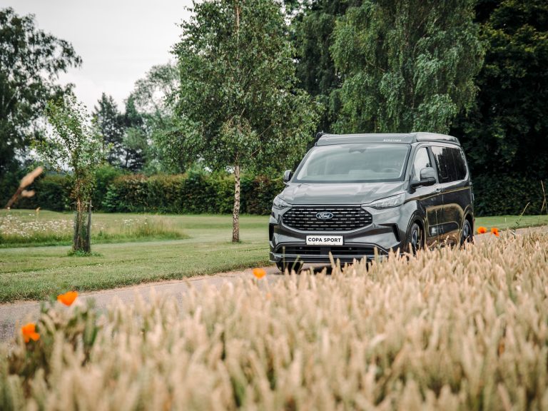 Ford Transit Camper mit geschlossenem Aufstelldach auf einem Parkweg – vor dem Wohnmobil reife Weizenfelder, im Hintergrund Zäune und dichter Wald. Idyllische Camping-Szene in ländlicher Natur.