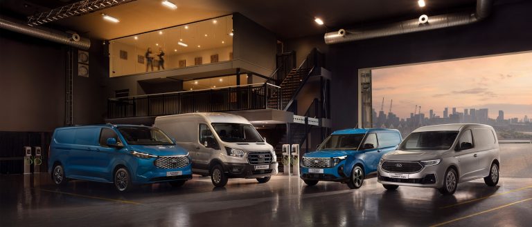 Vier Ford Transit-Modelle in unterschiedlichen Größen und Farben stehen in einer modernen Gewerbehalle mit mehreren Ladestationen für Elektrofahrzeuge. Im verschwommenen Hintergrund sind Hochhäuser einer Großstadt zu erkennen.