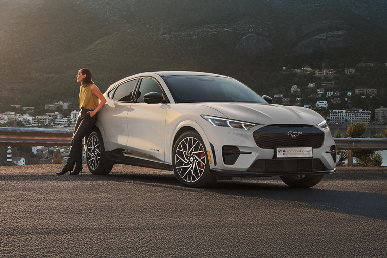 Eine Frau lehnt lässig an einem Ford Mustang Mach-E, der geparkt ist. Im Hintergrund sind moderne Gebäude und grüne Berge zu sehen. Das elektrische SUV-Coupé kombiniert sportliches Design mit urbaner Umgebung.